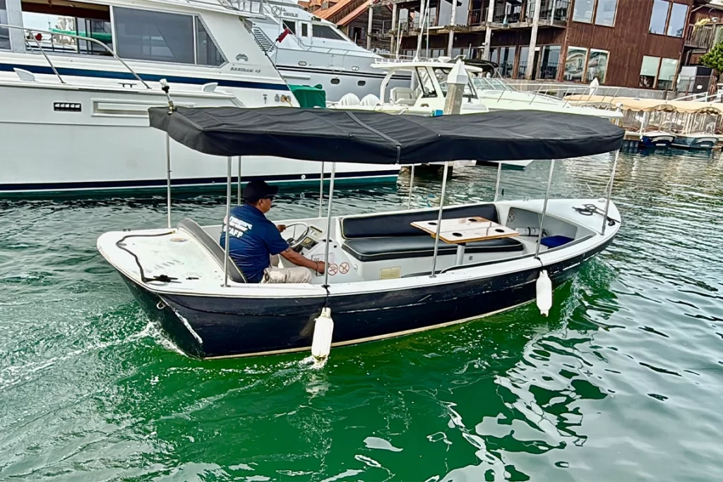A marina staff member steers a black canopy rental boat past parked luxury yachts.